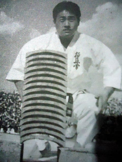 Masutatsu Oyama standing beside a tall stack of roof tiles before a stadium crowd.