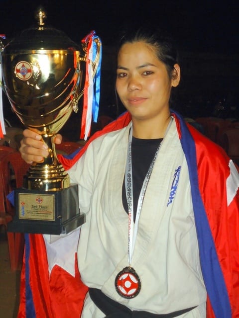 A Nepali karateka in a black belt holding a championship trophy, wearing a Kyokushin medal and draped in the national flag.